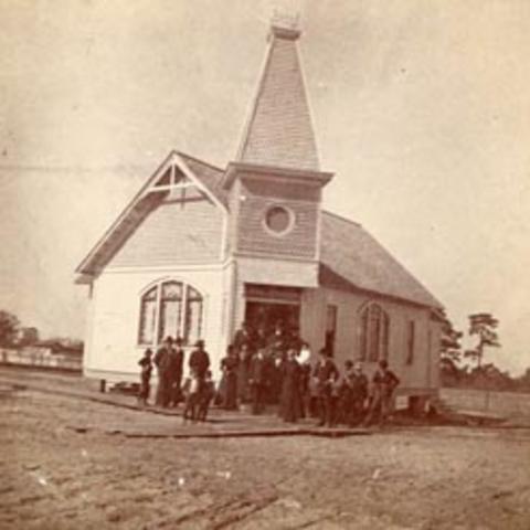 Sepia-toned photo of about 15 people standing in front of a small white church on bare ground.