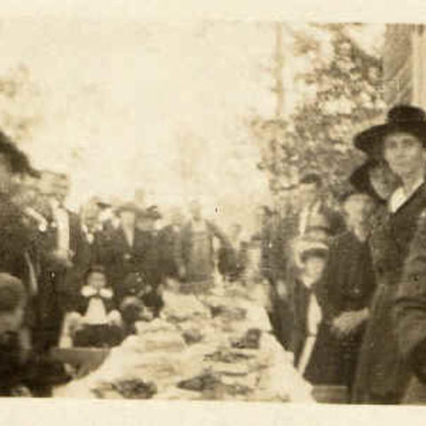 Sepia-toned photo of men, women, and children in church attire enjoying a meal at a long outdoor table